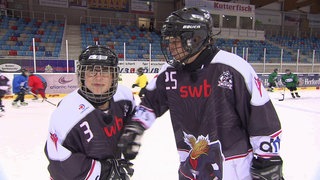 Zwei Personen stehen in Eishockeymontur in einer Eishalle. 